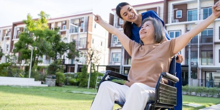 Happy Senior Woman Pushed in a Wheelchair by a Nurse Outside Independent Living Facility